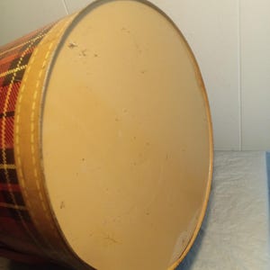 May include: A vintage, cylindrical container with a plaid pattern in red, yellow, and black. The lid is a light tan color, and the container has a tan band around the edge. The container is likely made of metal.