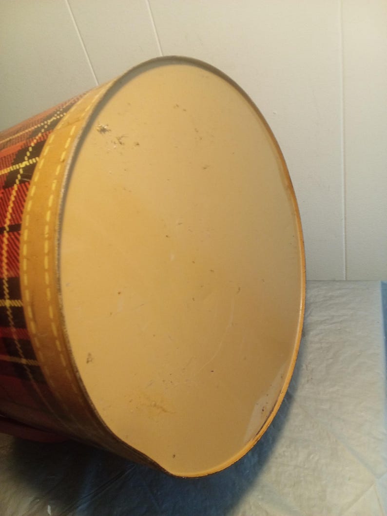 May include: A vintage, cylindrical container with a plaid pattern in red, yellow, and black. The lid is a light tan color, and the container has a tan band around the edge. The container is likely made of metal.