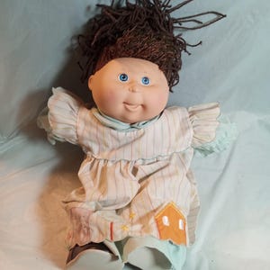 Puede incluir: Una muñeca con cabello de lana marrón oscuro y ojos azules, que viste un vestido claro con finas rayas verticales en tonos pastel. El vestido presenta mangas con volantes y un pequeño parche naranja en forma de casa con estrellas amarillas.