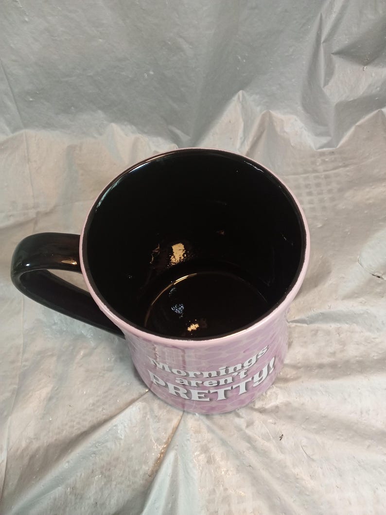 Puede incluir: Una taza de caf&eacute; de cer&aacute;mica con interior y asa negros. El exterior es de color lila claro con las palabras "mornings aren't PRETTY!" impresas en blanco. La taza est&aacute; sobre una superficie blanca arrugada.