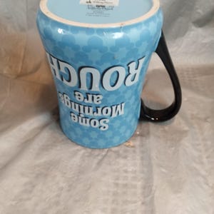 Pu&ograve; includere: Una tazza in ceramica azzurra con un manico nero. La tazza presenta un motivo di teste di Topolino e il testo "Some mornings are rough" in bianco. La tazza &egrave; un prodotto Disney.