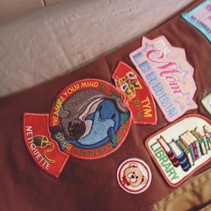 May include: A brown sash with various embroidered patches. Patches include the words "Mom Leader", "Library", "Treasure Your Mind", and "Netiquette". Other patches feature a dolphin, a teddy bear, and a tiger.