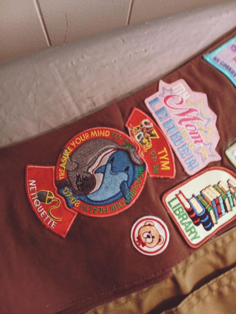 May include: A brown sash with various embroidered patches. Patches include the words "Mom Leader", "Library", "Treasure Your Mind", and "Netiquette". Other patches feature a dolphin, a teddy bear, and a tiger.