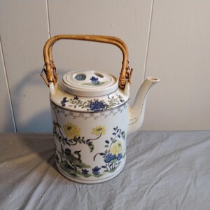 May include: A white porcelain teapot with a bamboo handle and floral design. The teapot has a lid with three holes and a spout. The floral design features yellow, blue, and green flowers.