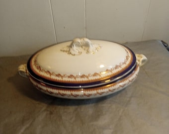 May include: Oval, lidded serving dish with a white base, adorned with a brown and blue decorative border. The lid features a decorative handle and gold accents. The dish has two side handles.