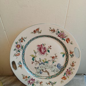 Peut inclure: Un plateau de service en céramique blanche avec un motif floral. Le plateau présente une fleur rose centrale avec des feuilles vertes et une bordure turquoise et verte. Des accents floraux roses, oranges et bleus ornent le bord. Deux poignées sont découpées.