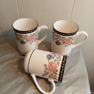 May include: Three white ceramic mugs with floral designs. Each mug features a black rim and handle, with a band of flowers in pink, blue, and purple. The mugs are arranged on a light gray surface.