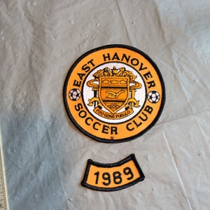 May include: Orange and black embroidered patch with the text "East Hanover Soccer Club" and a crest with the text "Pro Bono Publico". A smaller black and orange patch with the text "1989" is below the larger patch.
