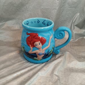 May include: A light blue ceramic mug featuring Ariel from The Little Mermaid. The mug has a curved handle and is decorated with images of Ariel, fish, and seaweed. The interior rim of the mug has a pattern of fish and stars.