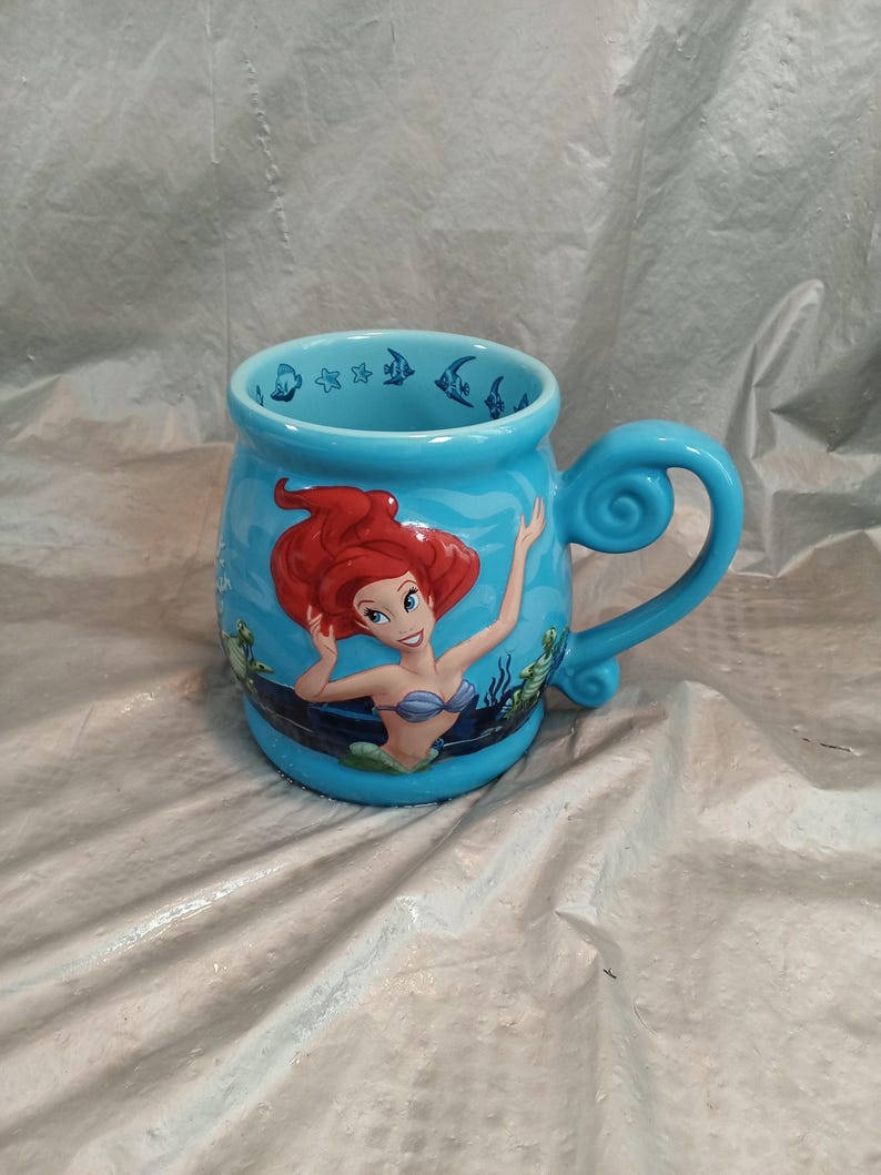 May include: A light blue ceramic mug featuring Ariel from The Little Mermaid. The mug has a curved handle and is decorated with images of Ariel, fish, and seaweed. The interior rim of the mug has a pattern of fish and stars.