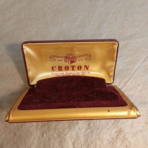 Puede incluir: Una caja de reloj vintage abierta con un interior de terciopelo burdeos y un borde dorado. La caja tiene el nombre "CROTON" impreso en el interior, junto con el texto "Cased and Timed in New York by THE CROTON WATCH CO."