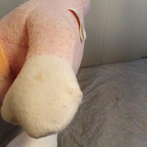 May include: A plush toy in shades of pink and white. The toy's body is a soft pink, with white accents on the feet. A small tag is attached to the side. The toy appears to be well-loved, with some discoloration.