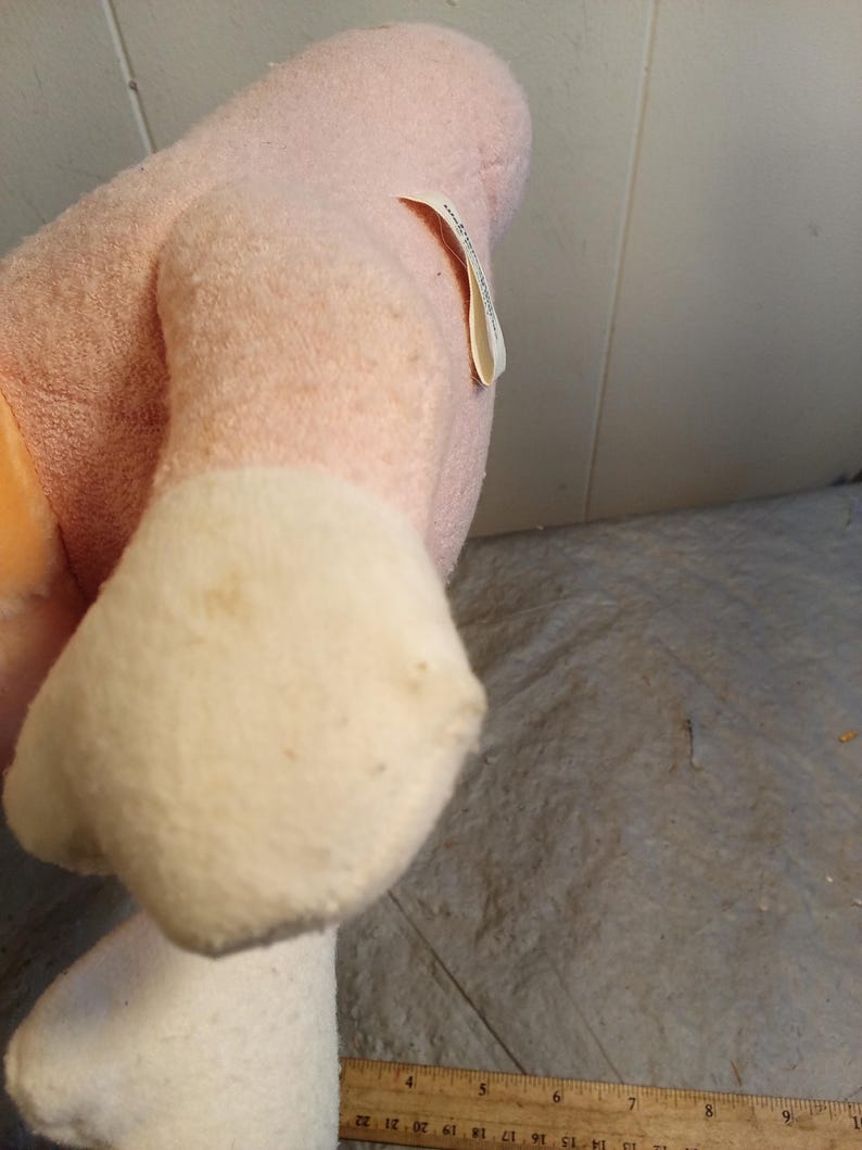 May include: A plush toy in shades of pink and white. The toy's body is a soft pink, with white accents on the feet. A small tag is attached to the side. The toy appears to be well-loved, with some discoloration.