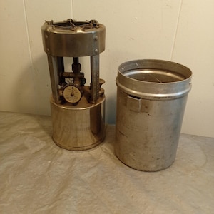 Pode incluir: Um fogão de campismo vintage prateado, com um depósito de combustível cilíndrico e um recipiente de metal separado. O fogão tem uma área de queimador circular e uma válvula de controlo. O metal tem um aspeto ligeiramente envelhecido.