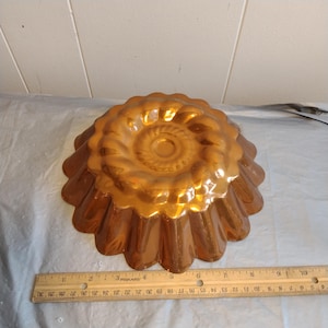 Op de afbeelding: Een koperkleurige, geribbelde, ronde cakevorm met een decoratief swirl-ontwerp in het midden. De vorm is ongeveer 20 cm in diameter.