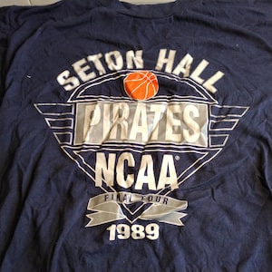 May include: A navy blue t-shirt with a silver and white graphic of the Seton Hall Pirates basketball team. The graphic includes the team name, a basketball, the NCAA logo, and the text "Final Four 1989".