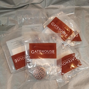 May include: Several clear plastic bags containing hardware components. Each bag has a red and white label with the text "GATEHOUSE - A STRONG IMPRESSION". One bag contains a copper-colored, circular knob with a patterned design.