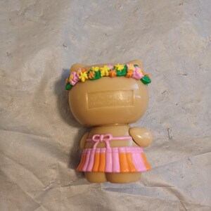 Op de afbeelding: Een bruin katachtig beeldje met een bloemenkrans en een roze en oranje rok. Het beeldje heeft kleine armen en een strikdetail in de taille. De bloemenkrans heeft gele, roze en groene bloemen.