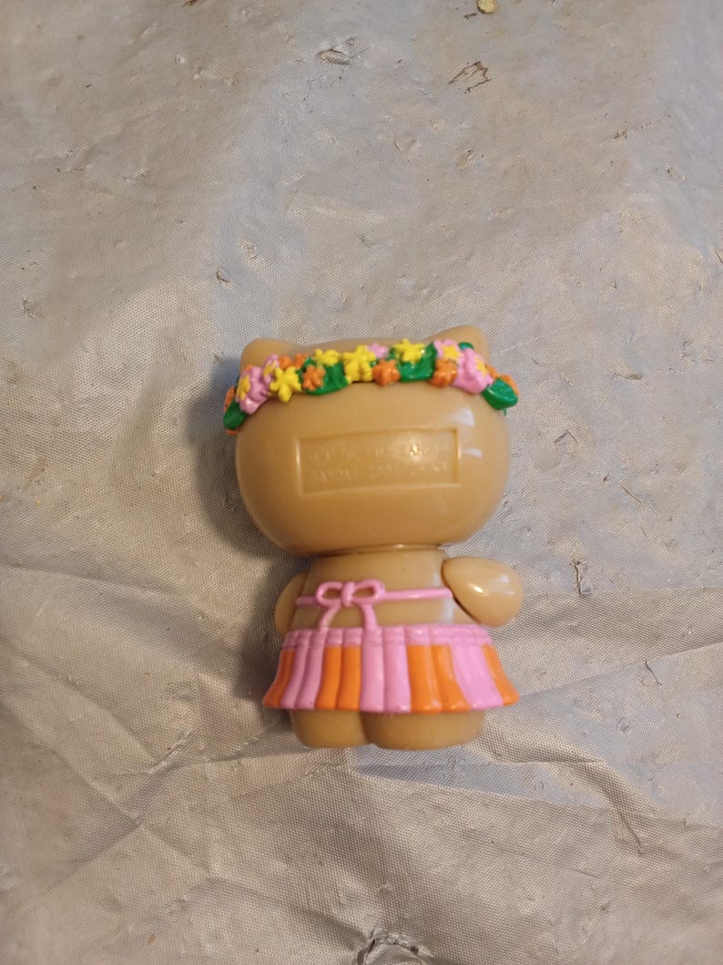 Op de afbeelding: Een bruin katachtig beeldje met een bloemenkrans en een roze en oranje rok. Het beeldje heeft kleine armen en een strikdetail in de taille. De bloemenkrans heeft gele, roze en groene bloemen.