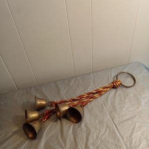 May include: A set of five brass bells hanging from a twisted gold and red rope, with a large metal ring at the top. The bells are arranged in a cascading pattern, creating a decorative and musical element.