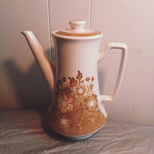 Puede incluir: Cafetera de cerámica blanca con tapa y asa. La cafetera presenta un diseño floral en tonos marrones y beige. El pico es largo y delgado, y el asa es rectangular.