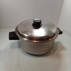 May include: A silver-toned metal cooking pot with a matching lid. The pot features two black rectangular side handles and a black knob on the center of the lid. The metal surface has a smooth, reflective finish, and the overall design is functional and simple.