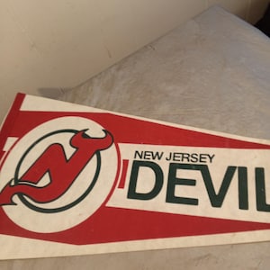 Puede incluir: Un banderín vintage de los New Jersey Devils. El banderín es blanco con rayas rojas y presenta el logotipo del equipo y las palabras "NEW JERSEY DEVILS" en verde. El logotipo es una "N" roja con una cabeza de diablo.