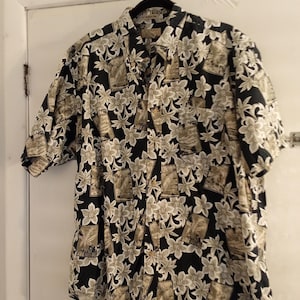 Puede incluir: Camisa de manga corta con fondo negro y estampado floral en tonos beige y blanco. La camisa está colgada de una percha negra contra una puerta blanca.