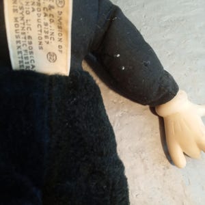 May include: A vintage Minnie Mouse plush doll, featuring a black outfit and a tag with text. The doll has a black body, a black sleeve, and a cream-colored hand. The tag reads "DIVISION OF ARRIE & CO. INC." and other text.