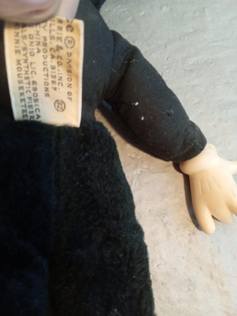 May include: A vintage Minnie Mouse plush doll, featuring a black outfit and a tag with text. The doll has a black body, a black sleeve, and a cream-colored hand. The tag reads "DIVISION OF ARRIE & CO. INC." and other text.