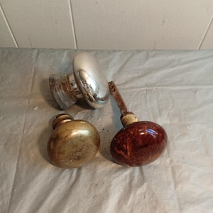 May include: Three antique door knobs. One is silver-colored with a tarnished base, another is gold-toned, and the third is a dark red-brown with a marbled appearance. The knobs are arranged on a white surface.