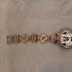 May include: A silver-toned wristwatch featuring a round face with a Mickey Mouse graphic, black numerals, and colourful hands. The metal link bracelet alternates between rectangular links embossed with the word MICKEY and circular links with icons.