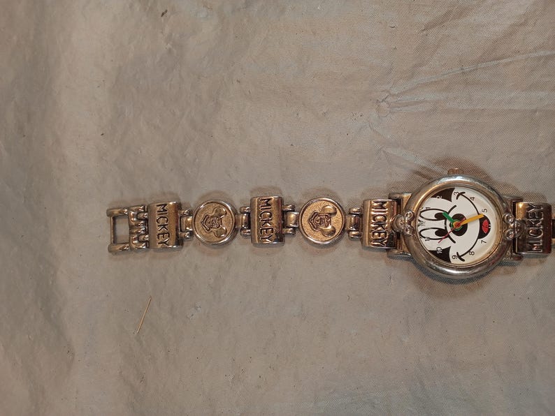 May include: A silver-toned wristwatch featuring a round face with a Mickey Mouse graphic, black numerals, and colourful hands. The metal link bracelet alternates between rectangular links embossed with the word MICKEY and circular links with icons.