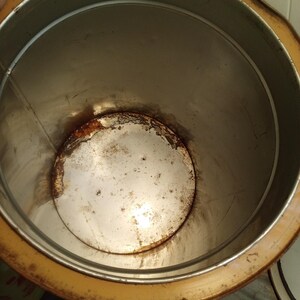 May include: An open, cylindrical metal container with a rusty interior and a yellow-brown rim. The inside of the container shows signs of age and wear, with discoloration and rust visible on the bottom and sides.