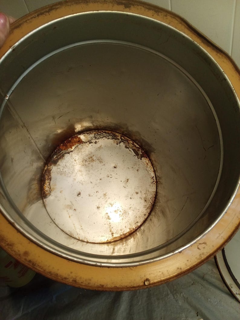May include: An open, cylindrical metal container with a rusty interior and a yellow-brown rim. The inside of the container shows signs of age and wear, with discoloration and rust visible on the bottom and sides.