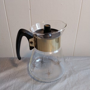 May include: A glass coffee pot with a gold band around the top and a black handle. The pot has a starburst design on the bottom.
