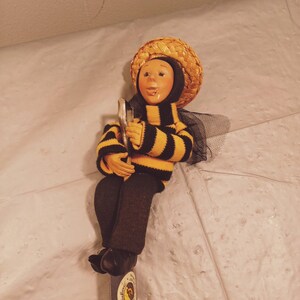May include: A yellow and black striped doll wearing a straw hat and holding a small metal object. The doll is sitting on a wooden snowboard with a yellow and black sticker that says "Schnitzler".