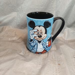 Pu&ograve; includere: Una tazza da caff&egrave; in ceramica con un'immagine a cartoni animati di Topolino. La tazza ha uno sfondo azzurro con un motivo, un manico nero e un interno nero. Topolino &egrave; raffigurato in accappatoio, con in mano una tazza di caff&egrave; e si tocca l'occhio.