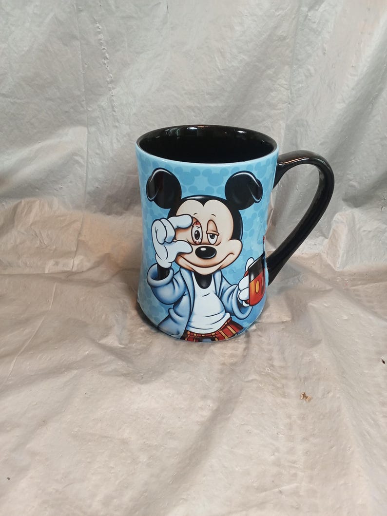 Pu&ograve; includere: Una tazza da caff&egrave; in ceramica con un'immagine a cartoni animati di Topolino. La tazza ha uno sfondo azzurro con un motivo, un manico nero e un interno nero. Topolino &egrave; raffigurato in accappatoio, con in mano una tazza di caff&egrave; e si tocca l'occhio.