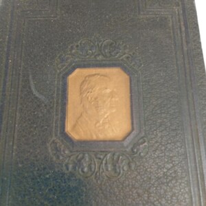 May include: Close-up of a vintage book cover. The cover is dark blue with a textured pattern. A gold-colored portrait is set within a decorative frame. Ornate corner designs add to the antique aesthetic.