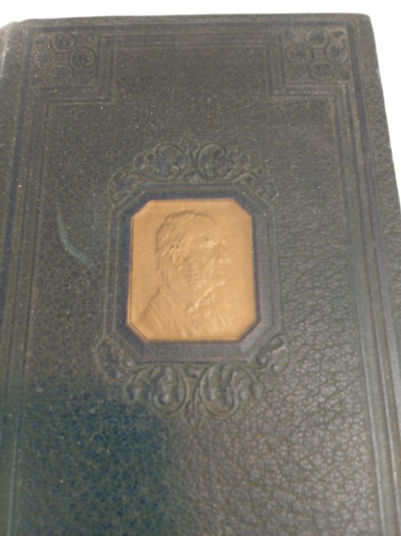 May include: Close-up of a vintage book cover. The cover is dark blue with a textured pattern. A gold-colored portrait is set within a decorative frame. Ornate corner designs add to the antique aesthetic.