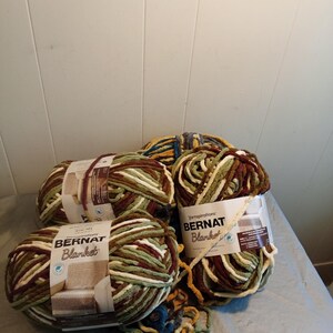 May include: Four skeins of Bernat Blanket yarn in various colors, including green, brown, cream, and burgundy. The yarn is thick and soft, ideal for blankets and other cozy projects. Each skein is labeled with the Bernat Blanket brand.