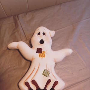 May include: A white ceramic ghost figurine with a surprised expression. The ghost has brown, yellow, and green fabric patches on its body.