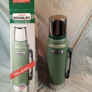 May include: A green Stanley Aladdin steel quart bottle with a chrome cap and a side handle, standing next to its original cardboard packaging. The box features the text Stanley, America's #1 Brand, and Steel Quart Bottle, with a capacity of 1 quart.