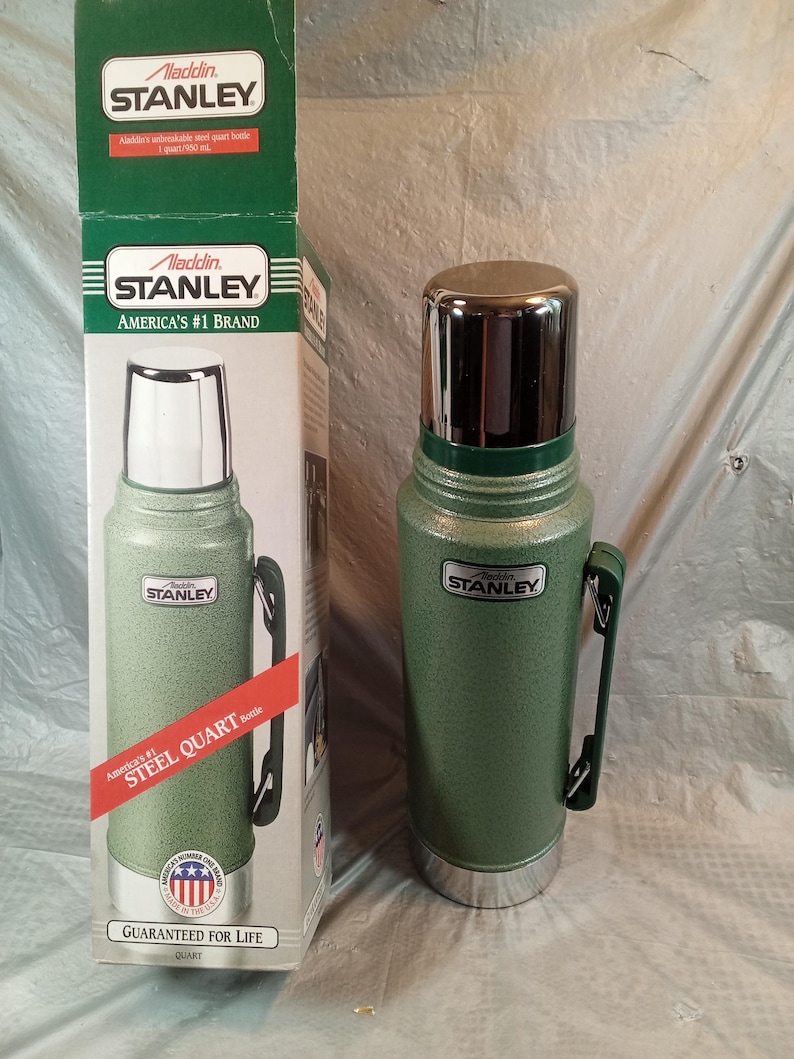 May include: A green Stanley Aladdin steel quart bottle with a chrome cap and a side handle, standing next to its original cardboard packaging. The box features the text Stanley, America's #1 Brand, and Steel Quart Bottle, with a capacity of 1 quart.