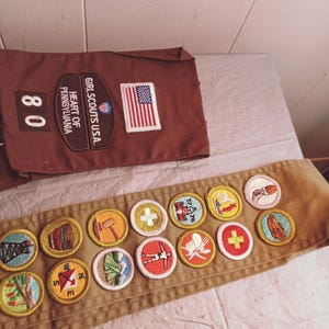May include: A brown Girl Scouts sash and a matching brown pocket with patches. The pocket has the text "GIRL SCOUTS U.S.A." and "HEART OF PENNSYLVANIA" with an American flag patch. The sash has several colorful embroidered patches.