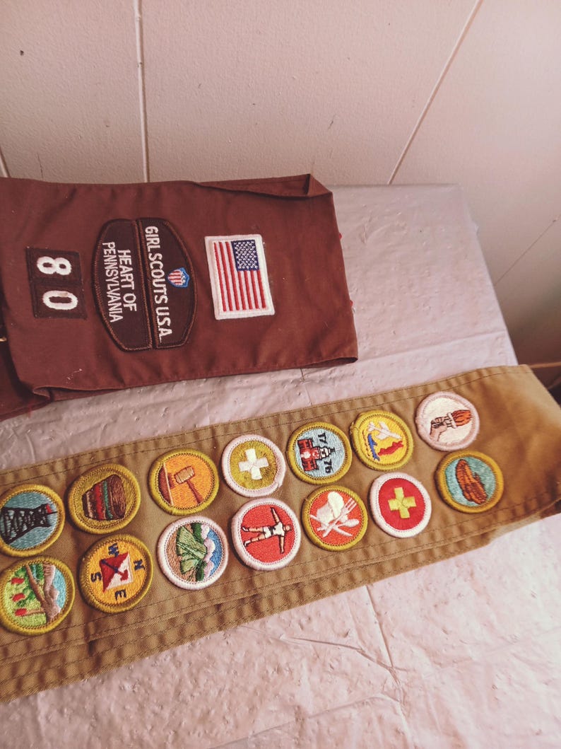 May include: A brown Girl Scouts sash and a matching brown pocket with patches. The pocket has the text "GIRL SCOUTS U.S.A." and "HEART OF PENNSYLVANIA" with an American flag patch. The sash has several colorful embroidered patches.