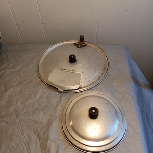 Pode incluir: Duas tampas de panela vintage de alumínio. Uma tampa tem uma alça e uma peça de metal articulada. Ambas as tampas têm um botão marrom escuro no centro. As tampas são circulares e têm um acabamento prateado.