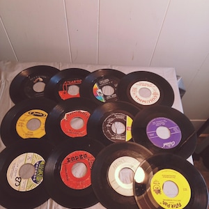 May include: A collection of 12 vintage vinyl records in various colours, including black, yellow, red, and white. The records are arranged in a circle on a white surface. Some of the records have text on them, including "Jamie", "Columbia", "Atlantic", and "Peter Pan".