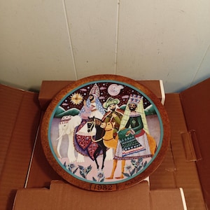 May include: A decorative plate featuring a colorful illustration of three figures riding animals, including an elephant and camels. The plate has a brown rim and a blue border. The year "1982" is printed at the bottom.
